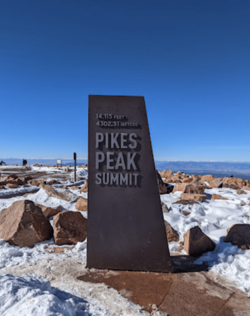 Sign at Pikes Peak Summit displaying elevation of 14,115 feet and snowy landscape under a clear blue sky.
