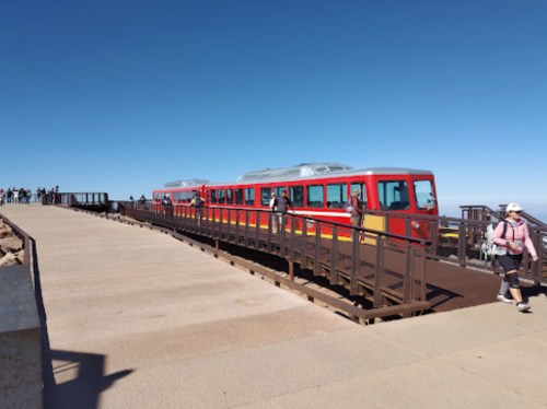 A red train on a platform with people walking nearby against a clear blue sky.