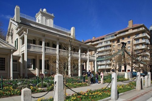 Historic mansion with a garden, surrounded by modern buildings and visitors enjoying the sunny day.
