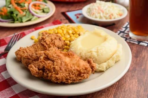 A plate of fried chicken with mashed potatoes, corn, and coleslaw, accompanied by a salad and a drink in the background.