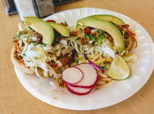 Two tostadas topped with shredded chicken, lettuce, tomatoes, avocado slices, and radishes, served with lime wedges.