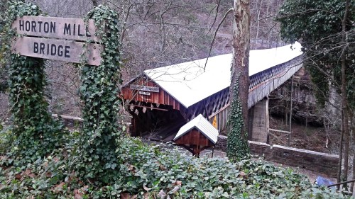 A wooden covered bridge surrounded by trees and greenery, with a sign reading "Horton Mill Bridge" nearby.