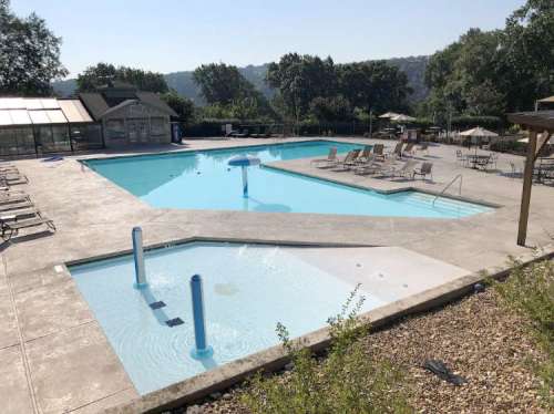 A sunny outdoor pool area featuring two pools, lounge chairs, and umbrellas surrounded by greenery.