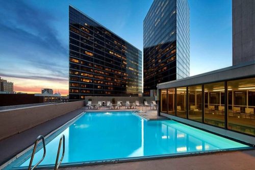 Rooftop pool with clear blue water, surrounded by modern glass buildings at sunset.