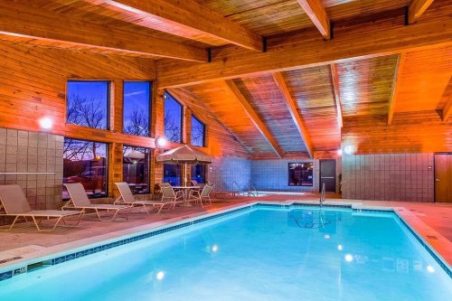 Indoor swimming pool with wooden beams, lounge chairs, and large windows reflecting evening light.