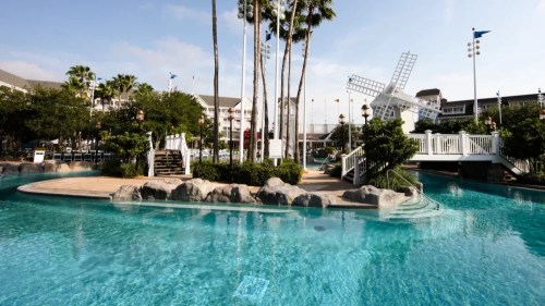A serene pool area surrounded by palm trees and a windmill, with clear blue water and a sunny sky.