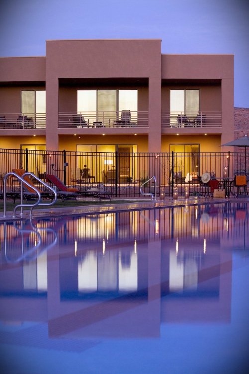 A tranquil pool reflects modern buildings with lit balconies, surrounded by lounge chairs and a serene evening sky.