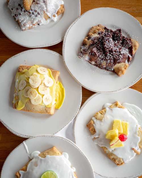 Five plates of dessert pastries topped with various fruits, cream, and drizzles, including banana, chocolate, and pineapple.