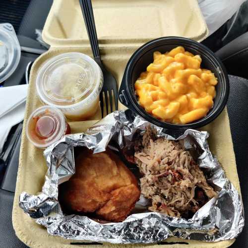 A takeout container with pulled pork, macaroni and cheese, a biscuit, and two small cups of sauce and fruit.