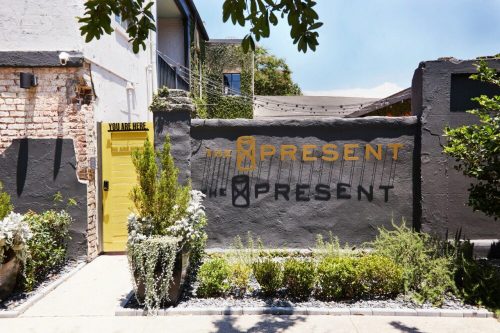 A colorful wall with "The Present" sign and a yellow door, surrounded by greenery and a clear blue sky.