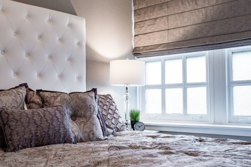 Cozy bedroom with a tufted white headboard, decorative pillows, a lamp, and a window with soft shades.