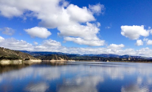 A serene lake reflects fluffy clouds and mountains under a bright blue sky.