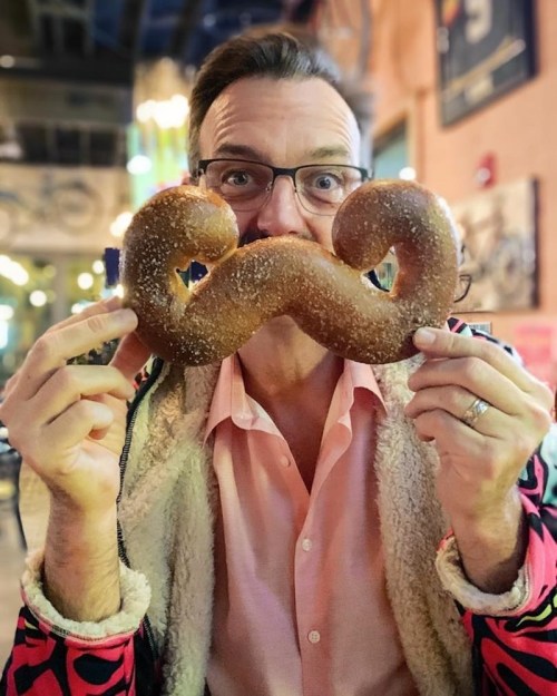 A man holds a large, pretzel-shaped mustache in front of his face, smiling in a colorful indoor setting.
