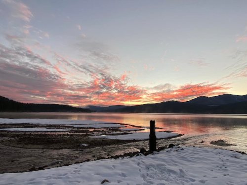 Sunset over a snowy landscape with a calm lake, mountains in the background, and colorful clouds in the sky.