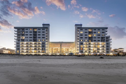 Two modern beachfront buildings with balconies, illuminated at dusk, facing a sandy beach under a colorful sky.