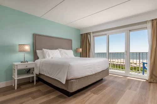 Cozy hotel room with a king bed, light blue walls, and a balcony overlooking the beach and ocean.