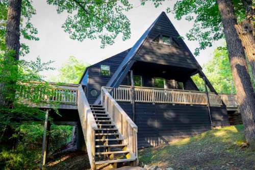 A black A-frame house surrounded by trees, featuring a wooden deck and stairs leading up to the entrance.