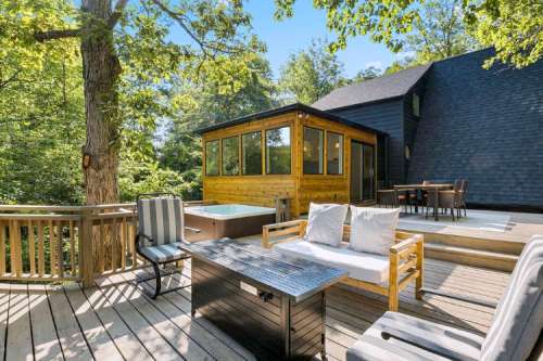 A wooden deck with seating, a hot tub, and a house surrounded by trees on a sunny day.