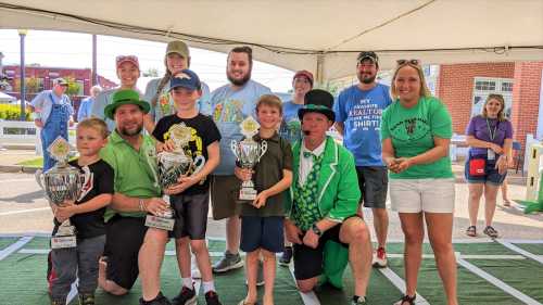 A group of people, including children and adults, celebrate with trophies under a tent at an outdoor event.