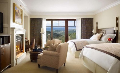 A cozy hotel room with two beds, a fireplace, and large windows showcasing a scenic view of mountains.