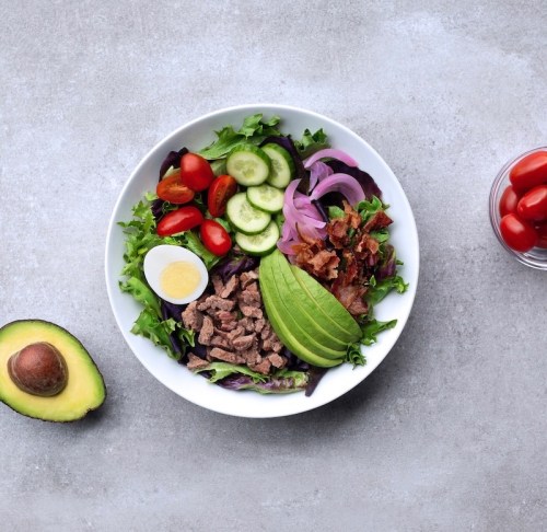 A colorful salad bowl with greens, cherry tomatoes, cucumber, avocado, beef, bacon, and a boiled egg, served with dressing.