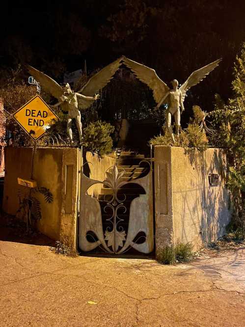 Two winged statues stand guard at a gated entrance, with a "Dead End" sign nearby, illuminated by night.