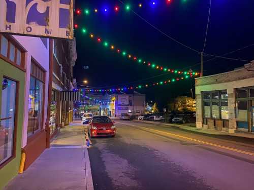 A quiet street at night adorned with colorful string lights, lined with shops and parked cars.