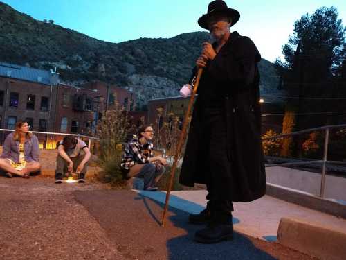 A person in a black cloak and hat stands with a cane, while others sit nearby in a dimly lit outdoor setting.