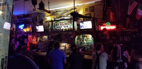 A lively bar scene with patrons gathered at the counter, surrounded by colorful lights and a variety of bottles on display.