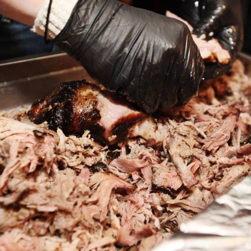 A close-up of hands in black gloves shredding tender, smoked meat on a tray.