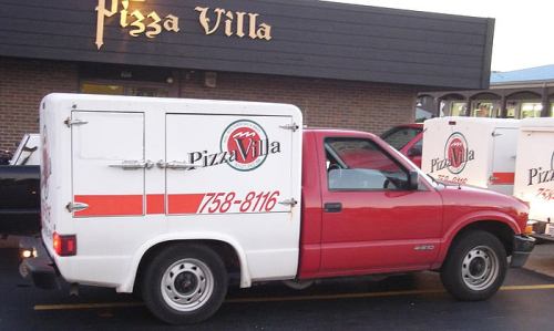A red delivery truck with a white cargo box, featuring the logo and contact number for Pizza Villa.