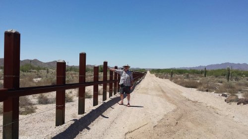 A person stands beside a tall border fence in a desert landscape under a clear blue sky.
