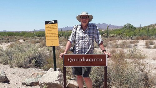 A person stands next to a sign that reads "Quitobaquito" in a desert landscape with cacti and mountains in the background.