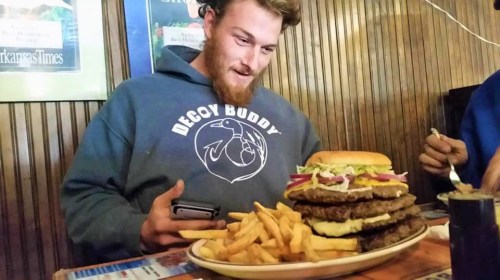 A man looks amazed at a towering burger with fries on a plate in front of him.