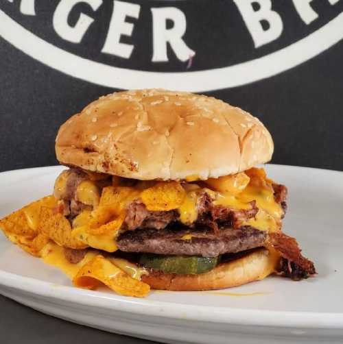 A hearty burger with two beef patties, melted cheese, Fritos, and pickles on a sesame seed bun, served on a plate.