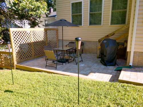 A patio area with a table, chairs, and a grill, surrounded by a wooden lattice and green grass.