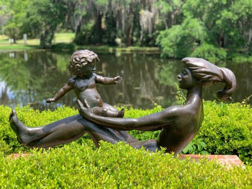 A bronze sculpture of a woman holding a child, surrounded by greenery with a pond in the background.