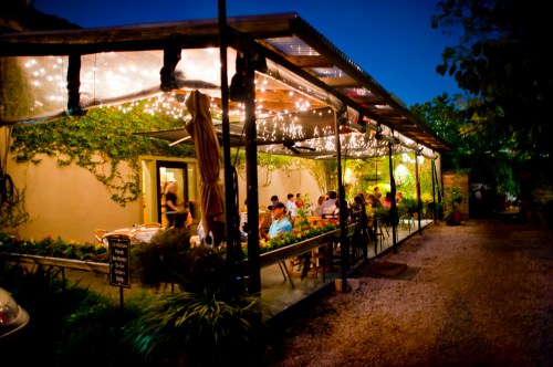 A cozy outdoor restaurant with string lights, surrounded by greenery, filled with diners enjoying the evening.