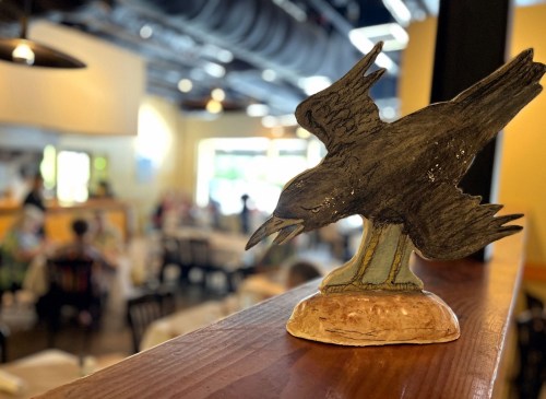 A handcrafted black bird figurine perched on a wooden ledge, with a restaurant scene blurred in the background.