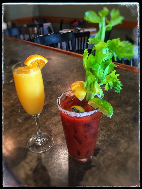 A glass of orange mimosa and a Bloody Mary garnished with celery and citrus slices on a bar counter.