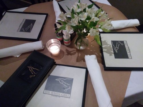 Table setting at The Raven Grill with menus, a candle, and a vase of flowers.