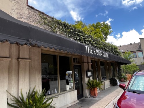 Exterior of The Raven Grill with a black awning, greenery, and a clear blue sky in the background.