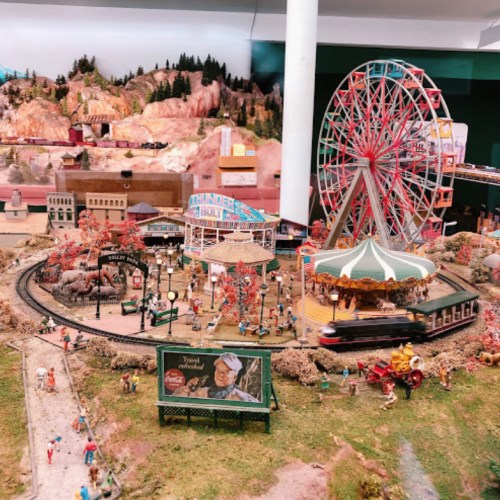 A detailed miniature amusement park scene with a carousel, Ferris wheel, and train, surrounded by trees and figures.