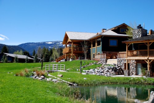 A scenic view of a rustic lodge with a stone facade, surrounded by green grass, a pond, and mountains in the background.