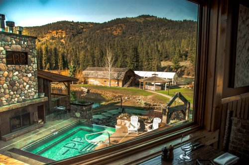 View from a cabin window showcasing a hot tub, rustic buildings, and a scenic mountain landscape.