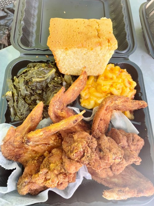 A takeout container with fried chicken wings, macaroni and cheese, collard greens, and a piece of cornbread.