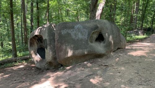 A large, weathered rock with two holes, surrounded by trees along a dirt path in a forested area.