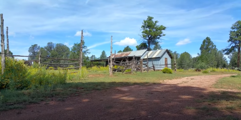 A rustic, weathered barn surrounded by trees and wildflowers under a blue sky with scattered clouds.