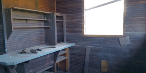 Interior of a rustic room with wooden walls, a workbench, shelves, and a window letting in natural light. Dusty surfaces.