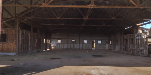 Interior of a large, empty, rustic building with wooden beams and weathered walls, illuminated by natural light.